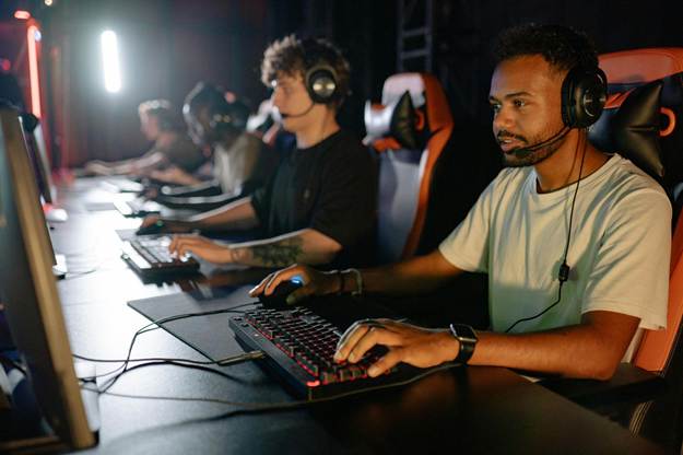 Gamers wearing headsets and focusing on playing a video game on desktop computers in a dimly lit gaming room.